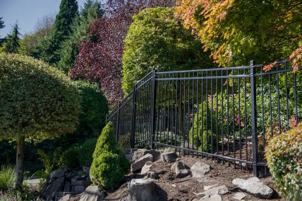 Black ornamental metal security fence panels installed on a sloped landscape with rock retaining walls and lush greenery.