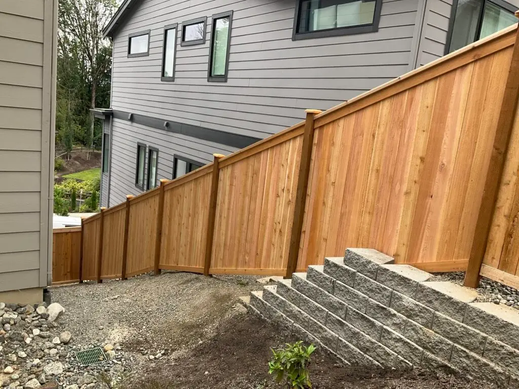 A racked fence on uneven ground angling downward to follow the terrain next to a gray stone retaining wall and modern home.
