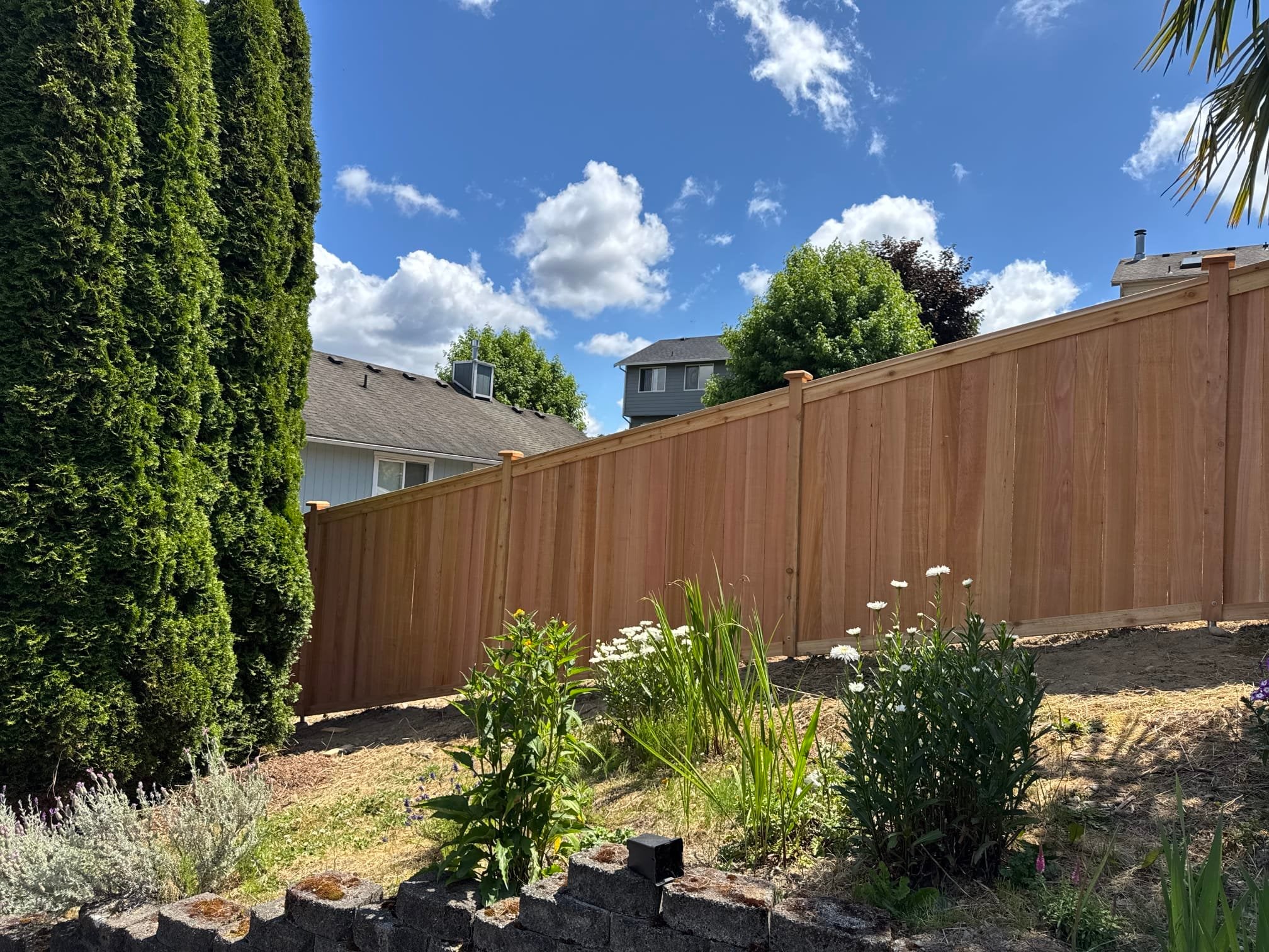A high-quality privacy fence on uneven ground with no visible gaps at the base, framed by green arborvitae and garden flowers.