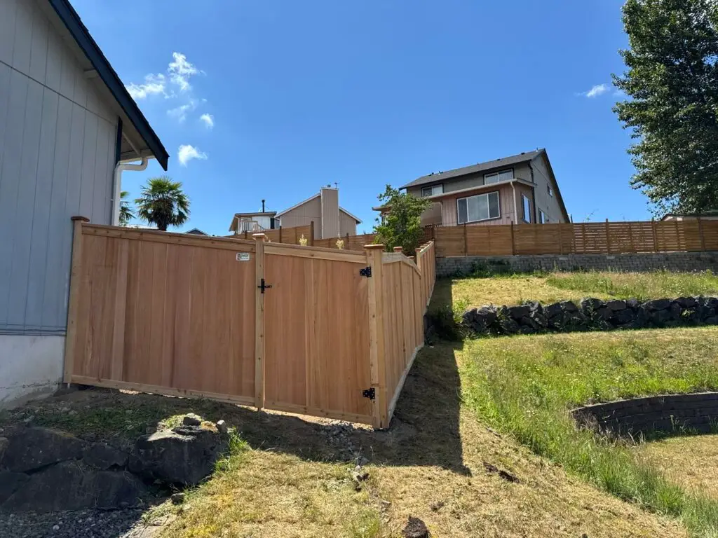 A wooden privacy fence with a gate installed along an uneven rocky slope near a house.