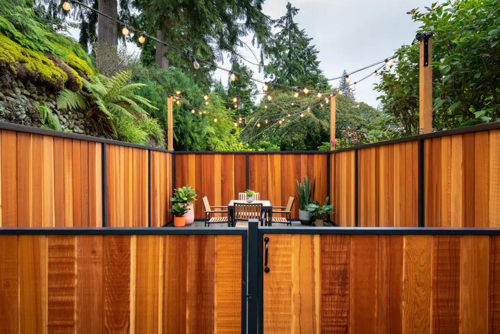 A private outdoor patio with a dining table and chairs enclosed by a premium vertical cedar wood fence with a sleek black frame, illuminated by overhead string lights.