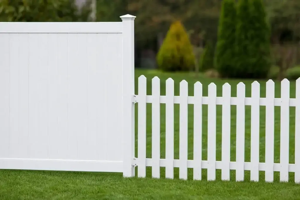 A side-by-side comparison of a tall white vinyl privacy fence panel and a shorter white vinyl picket fence section in a green backyard.