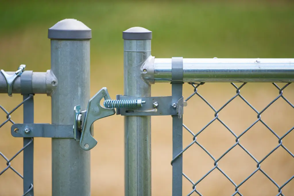 Close-up of standard chain link fence gate hardware, including galvanized steel latch, gate hinges, and tension bands.