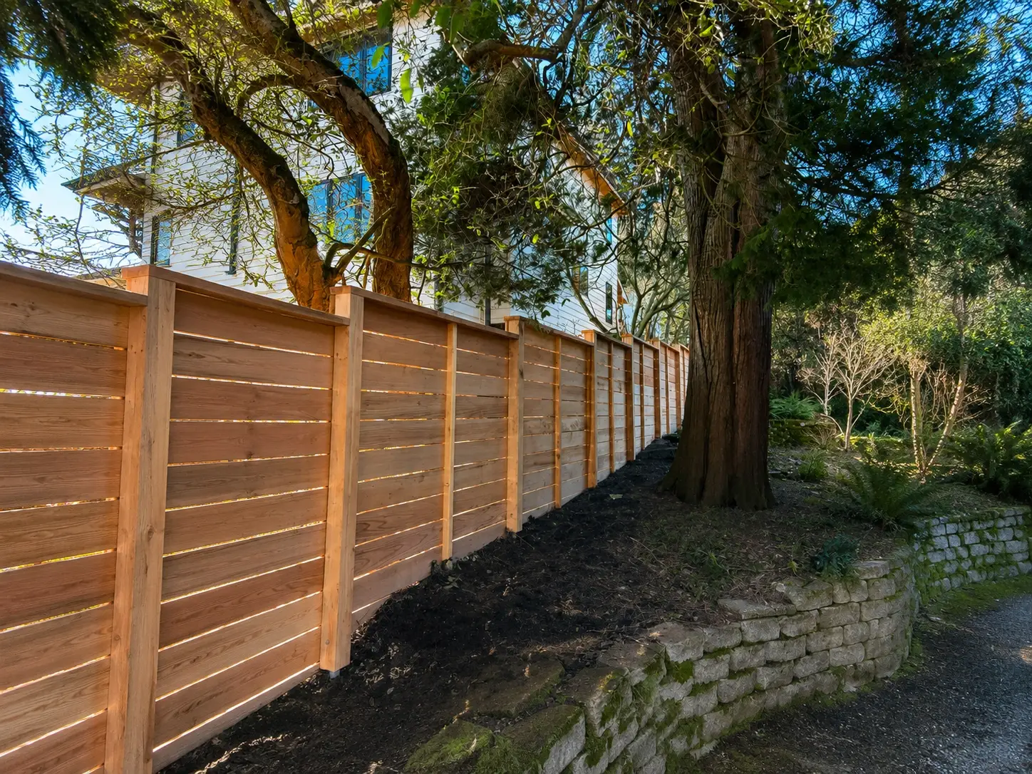 A simple wood fence design built along a sloped garden bed beside mature trees and a stone wall.